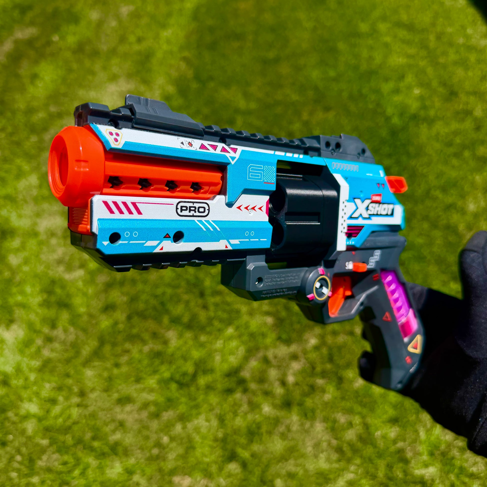A toy blaster pistol held by a hand against a green grass background. The blaster is primarily blue, white, and black with bright orange accents on the barrel and trigger, and purple details on the grip.