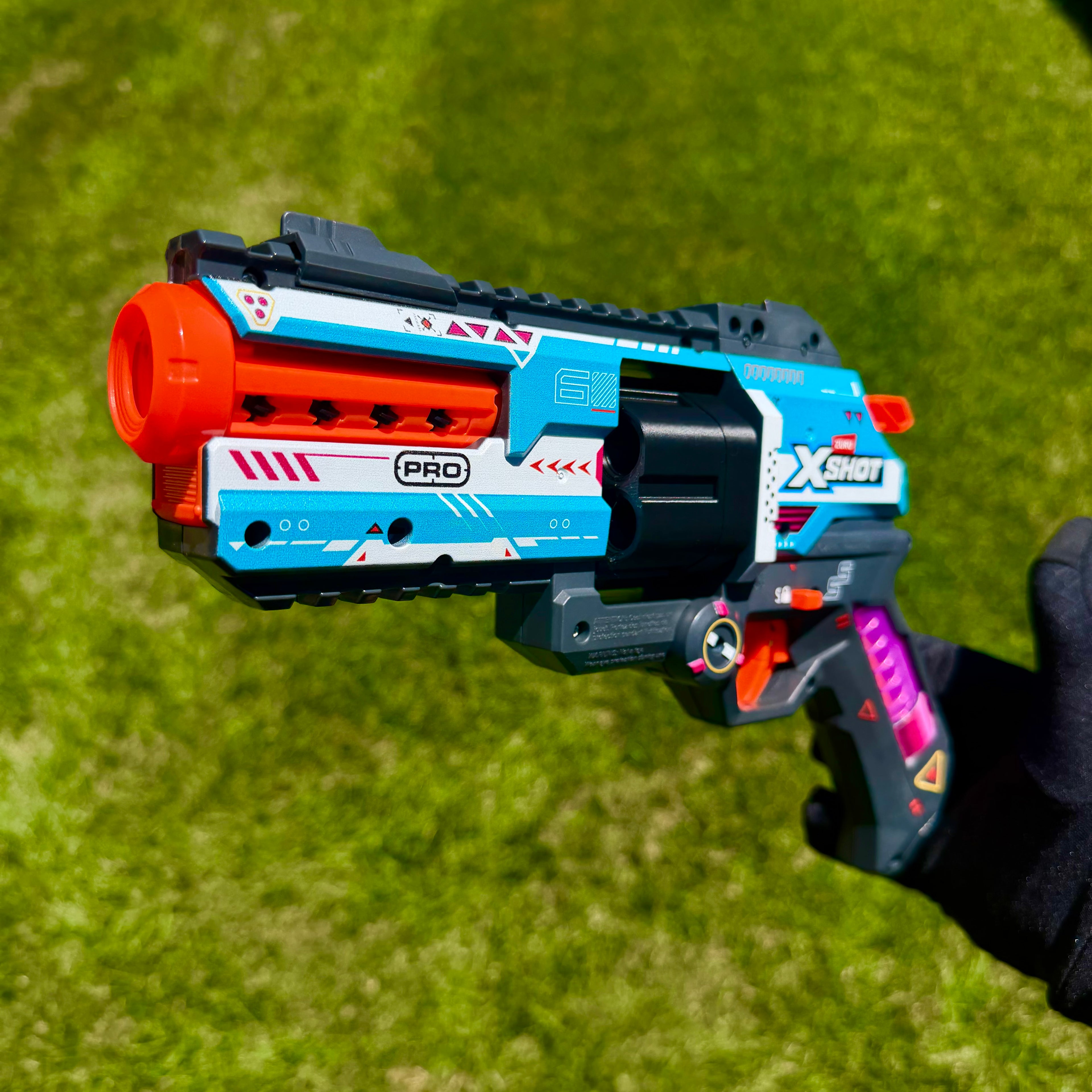 A toy blaster pistol held by a hand against a green grass background. The blaster is primarily blue, white, and black with bright orange accents on the barrel and trigger, and purple details on the grip.