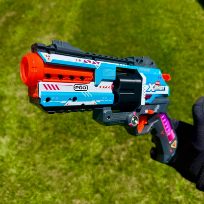 A toy blaster pistol held by a hand against a green grass background. The blaster is primarily blue, white, and black with bright orange accents on the barrel and trigger, and purple details on the grip.