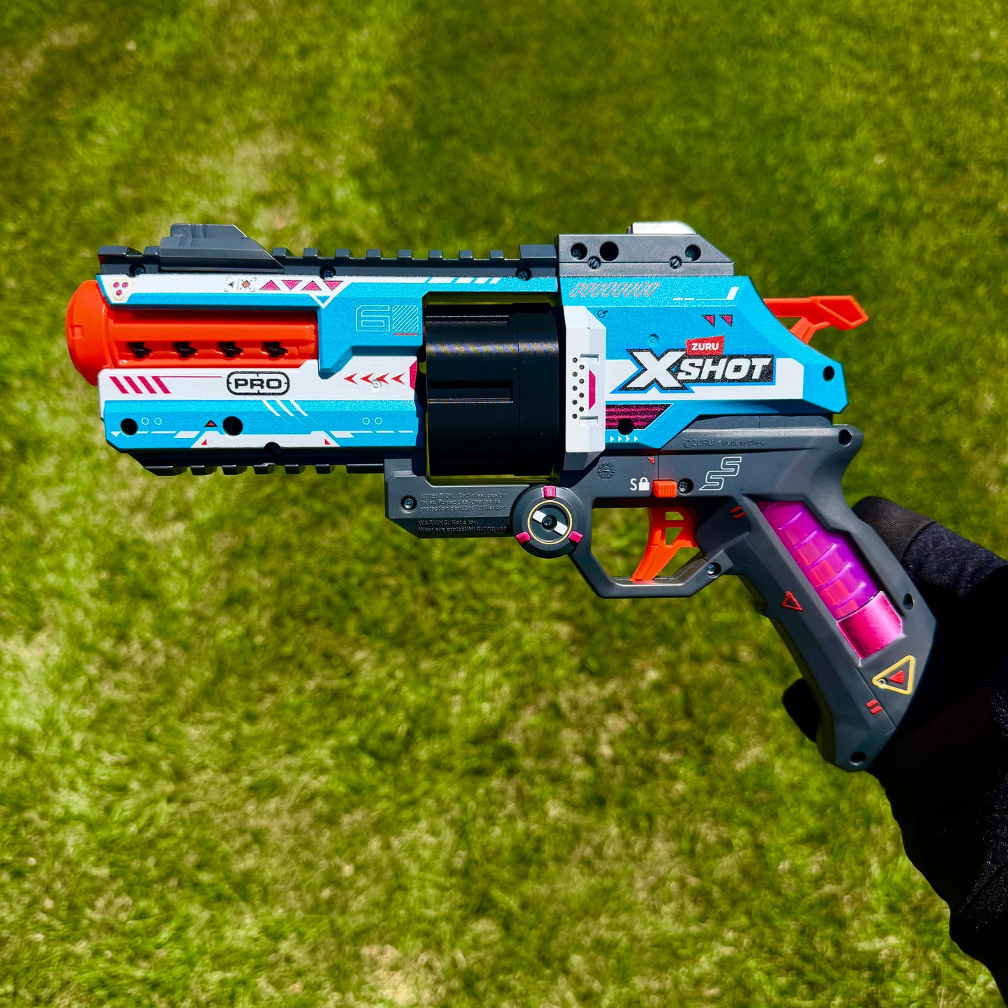 A toy blaster pistol held by a hand against a green grass background. The blaster features a blue, white, and black body with a bright orange muzzle, a black center barrel, and a pink magazine.